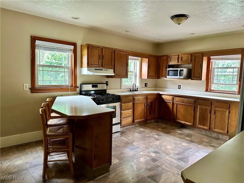 Kitchen with stainless steel appliances, under cabinet range hood, a textured ceiling, brown cabinetry, and healthy amount of natural light