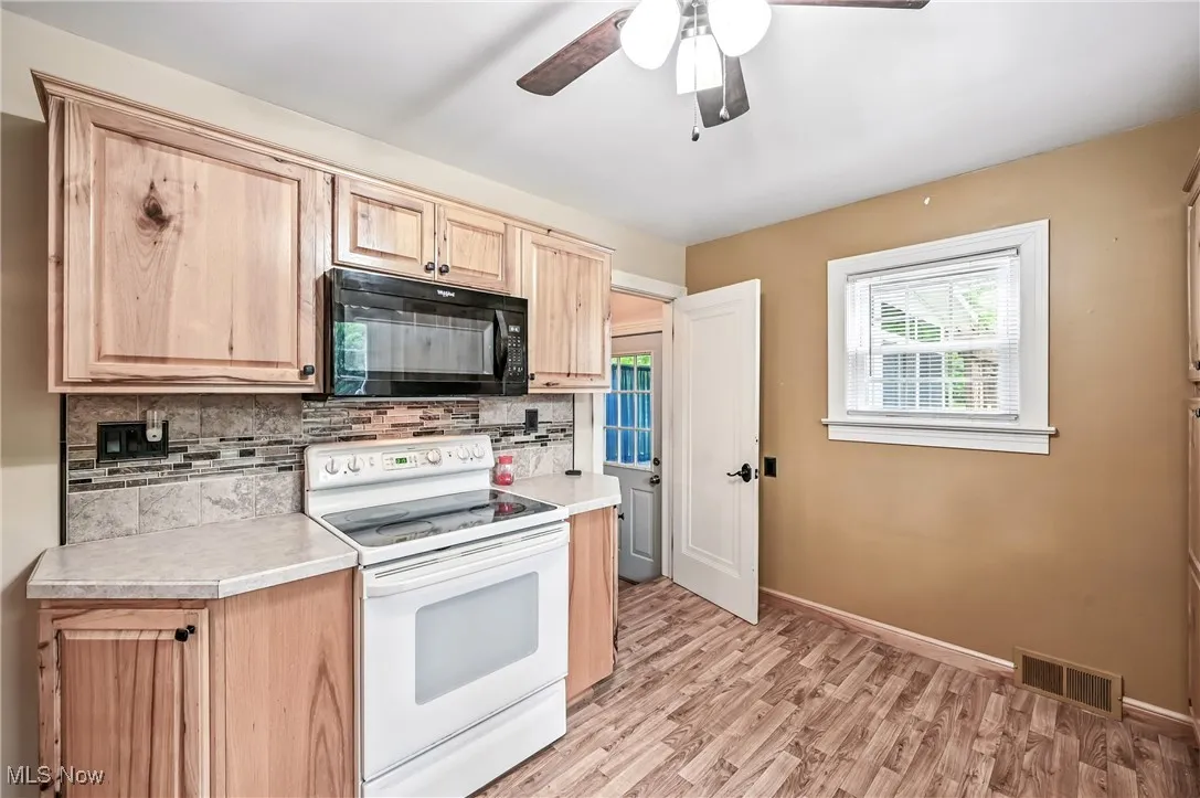 Kitchen with black microwave, white electric stove, light brown cabinetry, backsplash, and light countertops