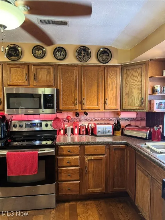 Kitchen featuring appliances with stainless steel finishes, tasteful backsplash, brown cabinetry, dark wood-style floors, and dark countertops