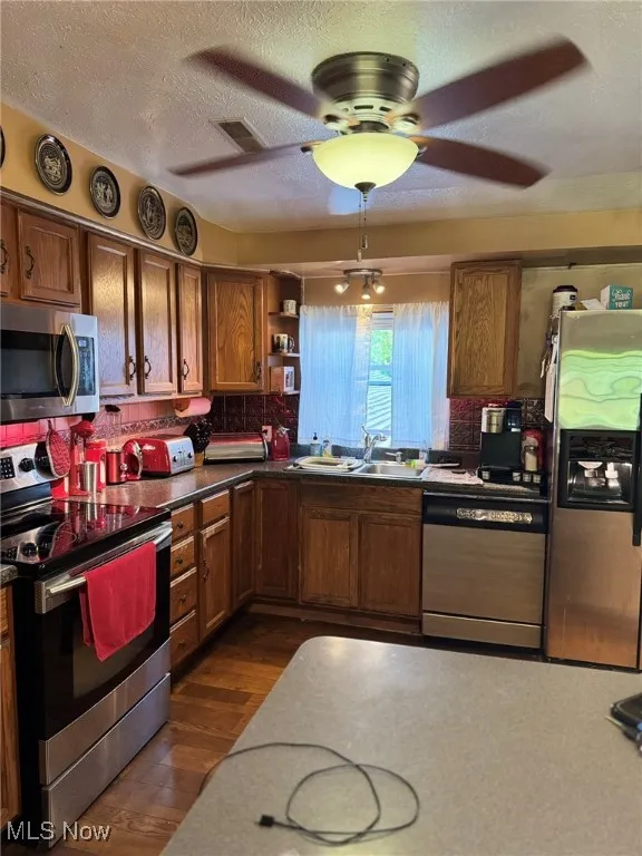 Kitchen with stainless steel appliances, brown cabinetry, ceiling fan, dark wood-style flooring, and tasteful backsplash