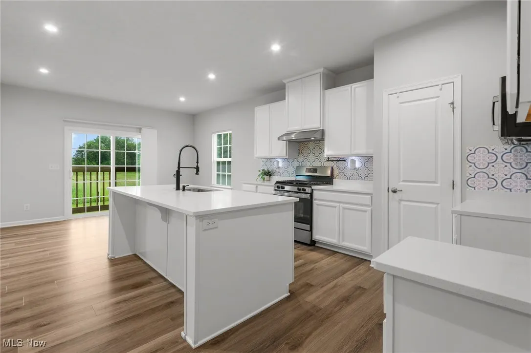 Kitchen featuring stainless steel gas stove, under cabinet range hood, backsplash, a center island with sink, and white cabinets