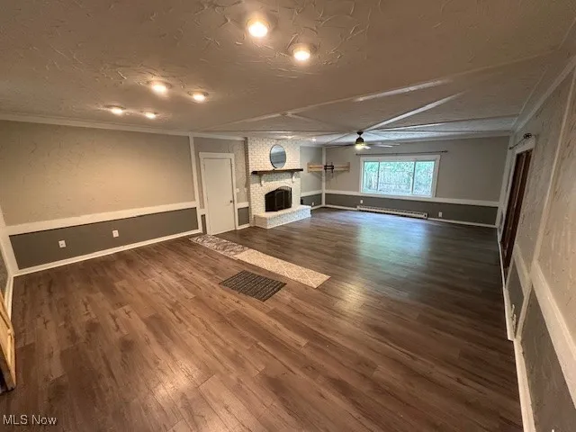 Unfurnished living room featuring a brick fireplace, dark wood-type flooring, ornamental molding, ceiling fan, and baseboard heating
