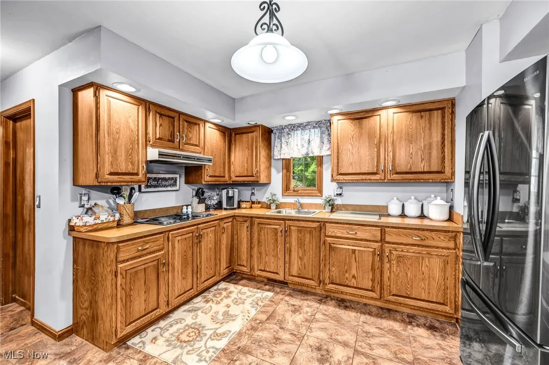 Kitchen with black appliances, under cabinet range hood, brown cabinetry, light countertops, and hanging light fixtures