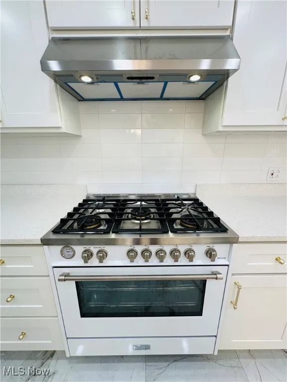 Kitchen with high end Italian range with gas cooktop, under cabinet range hood, light marble finish flooring, white cabinets, and decorative backsplash