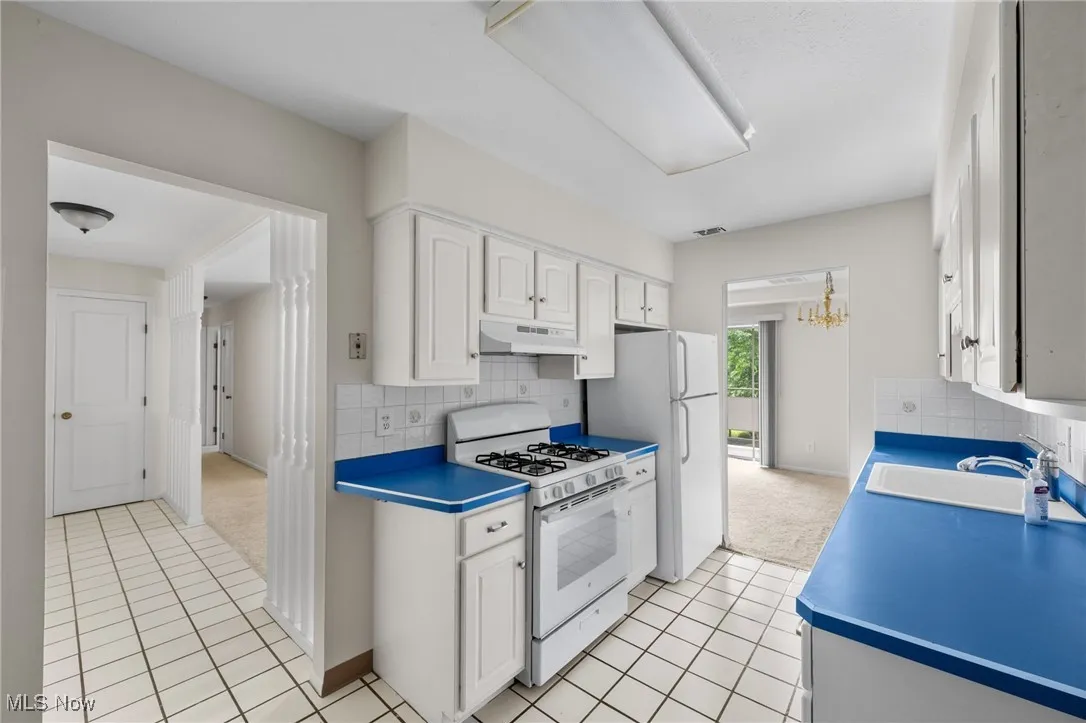 Kitchen featuring white appliances, under cabinet range hood, white cabinetry, light tile patterned floors, and decorative backsplash