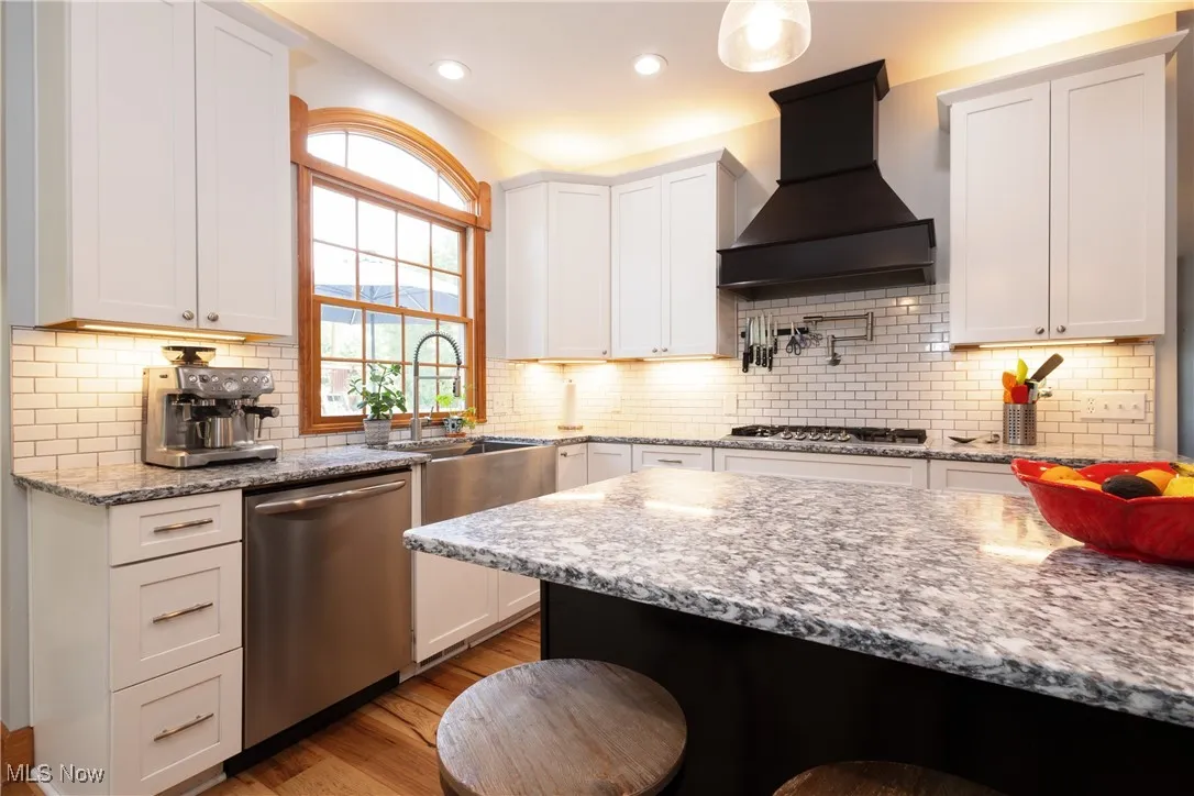 Kitchen with stainless steel appliances, premium range hood, light wood-style floors, white cabinets, and tasteful backsplash