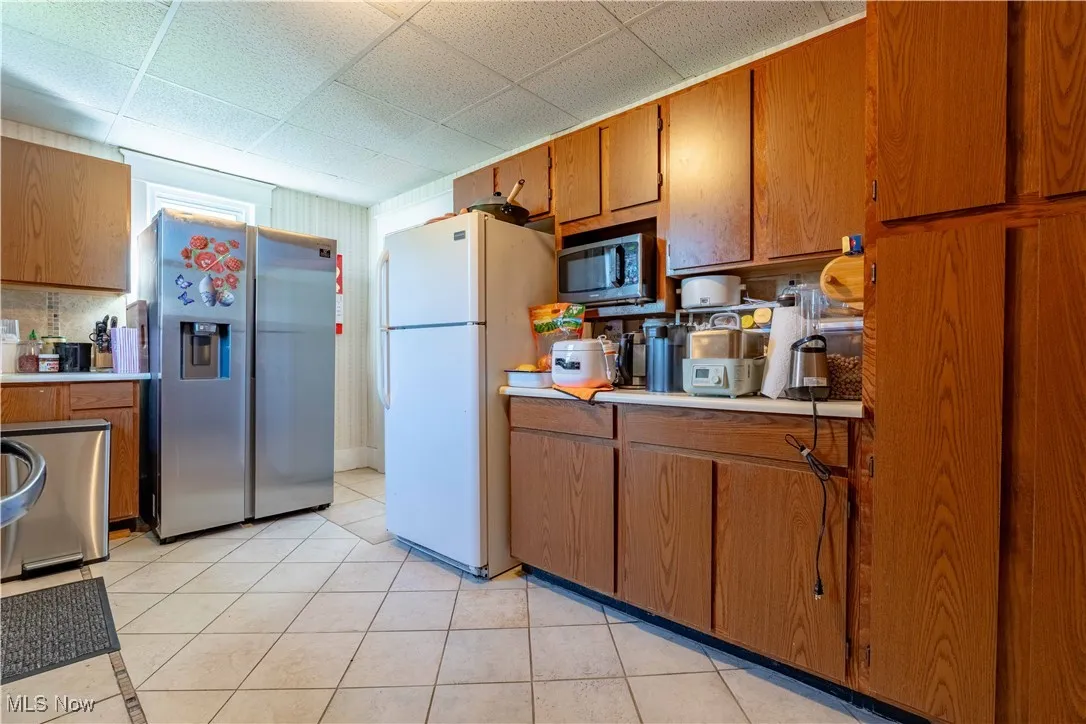Kitchen featuring stainless steel appliances, brown cabinets, light countertops, light tile patterned floors, and a drop ceiling