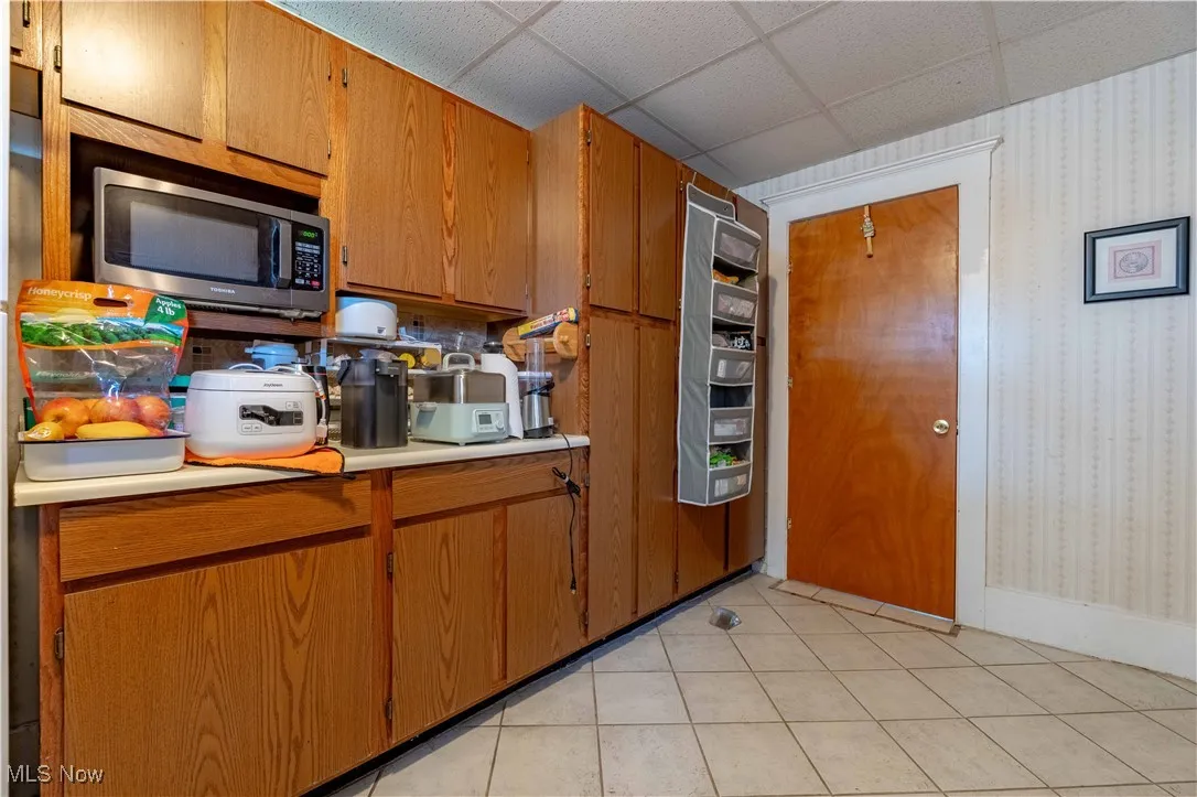 Kitchen featuring stainless steel microwave, light countertops, wallpapered walls, brown cabinets, and light tile patterned floors