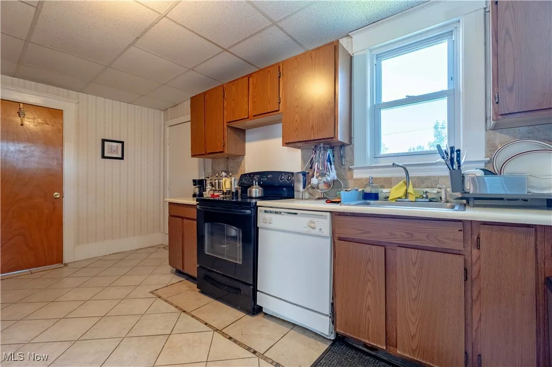 Kitchen with dishwasher, black range with electric cooktop, light countertops, light tile patterned flooring, and brown cabinetry