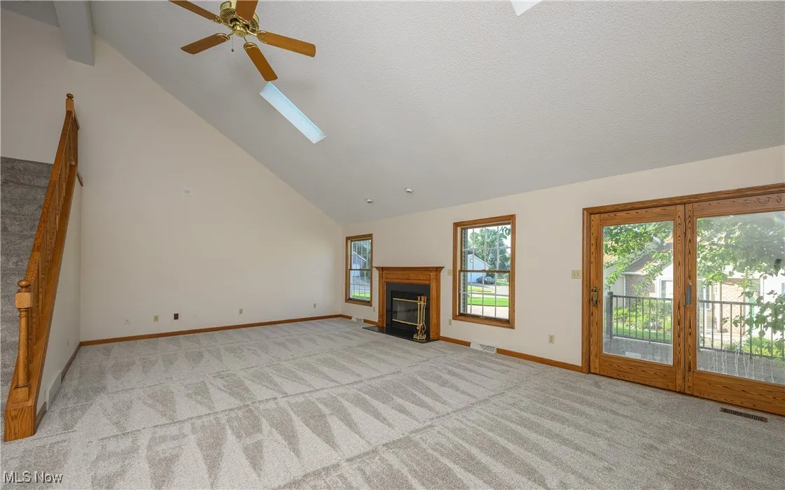 Unfurnished living room featuring a skylight, light colored carpet, a ceiling fan, high vaulted ceiling, and a fireplace with flush hearth