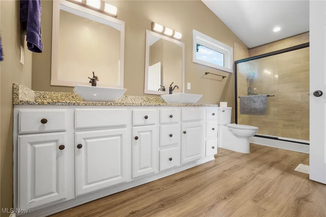 Bathroom with double vanity, a stall shower, wood finished floors, a baseboard heating unit, and vaulted ceiling