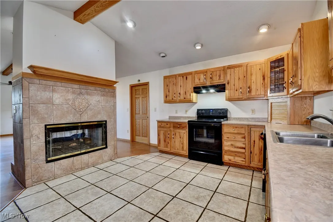 Kitchen with black electric range oven, under cabinet range hood, light countertops, a tile fireplace, and glass insert cabinets