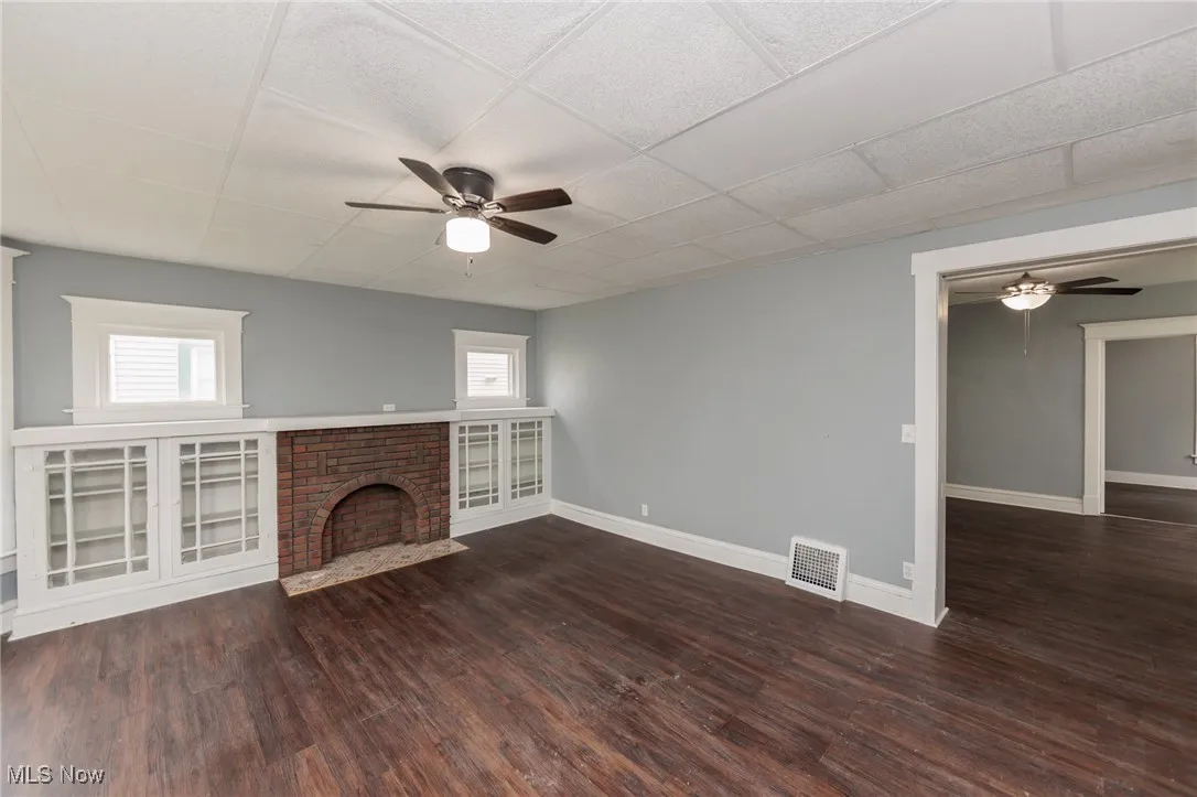 Unfurnished living room with a ceiling fan, plenty of natural light, wood finished floors, a brick fireplace, and a drop ceiling