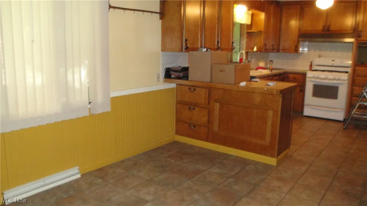 Kitchen featuring white gas range oven, a baseboard radiator, brown cabinetry, a peninsula, and tasteful backsplash