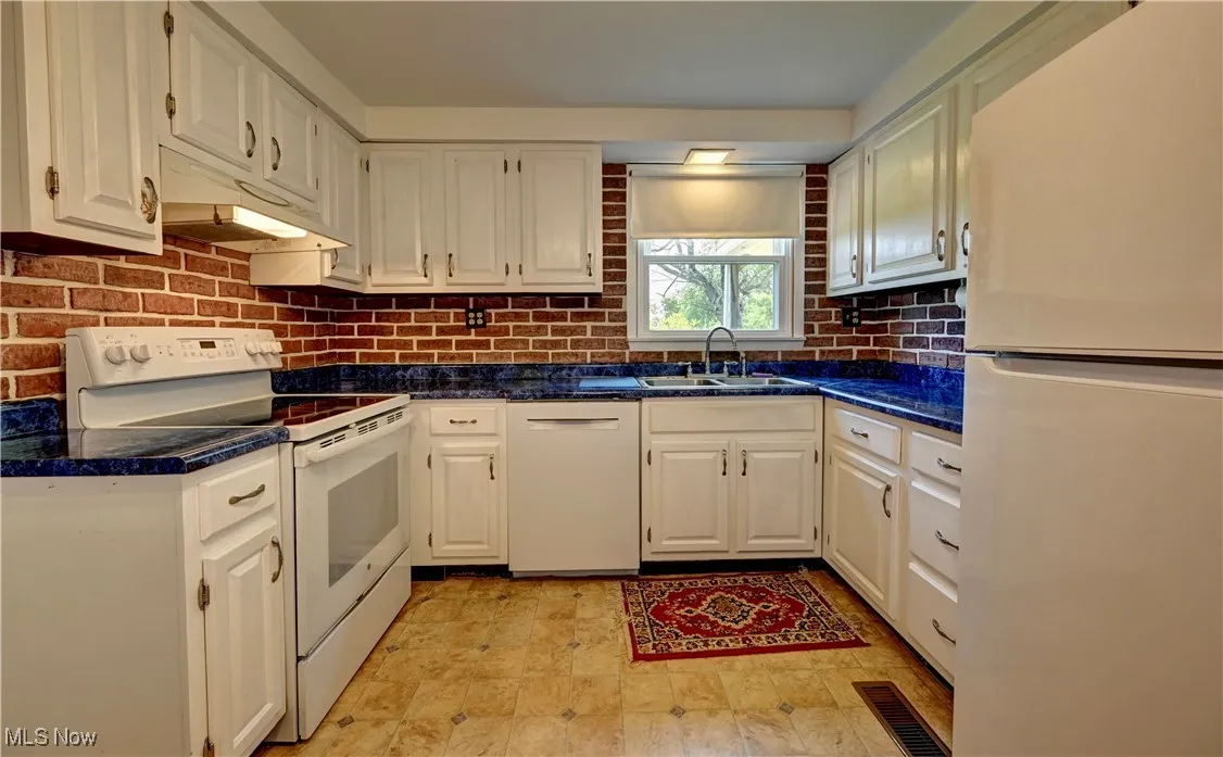 Kitchen with white appliances, white cabinetry, dark countertops, tasteful backsplash, and under cabinet range hood