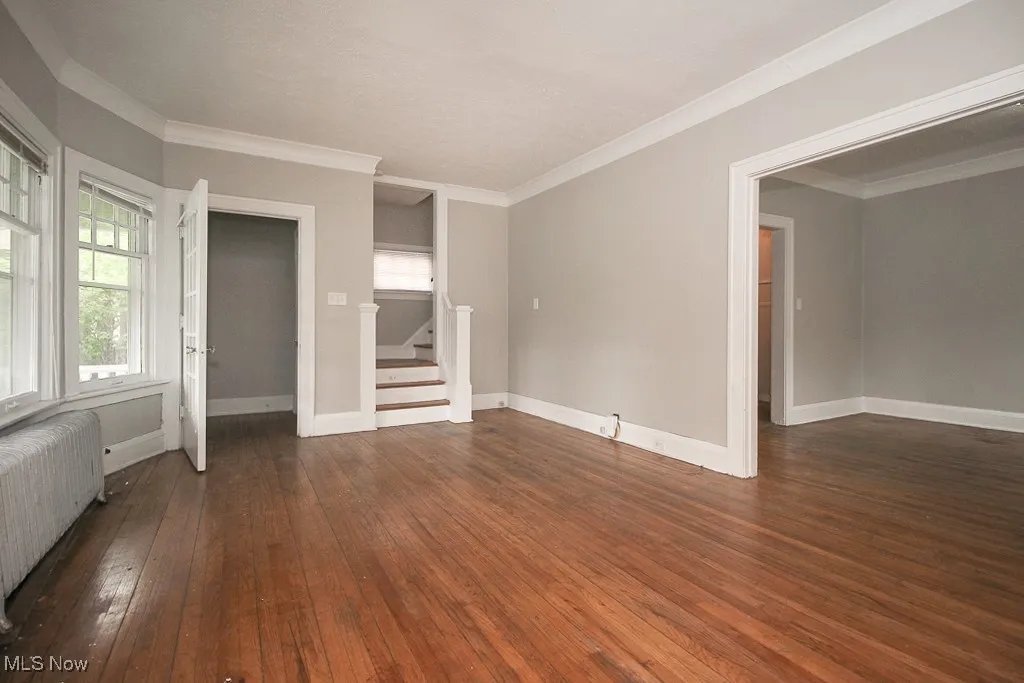 Empty room featuring radiator heating unit, stairs, wood-type flooring, and crown molding
