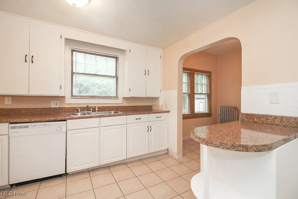 Kitchen featuring dishwasher, arched walkways, white cabinetry, light tile patterned flooring, and dark countertops