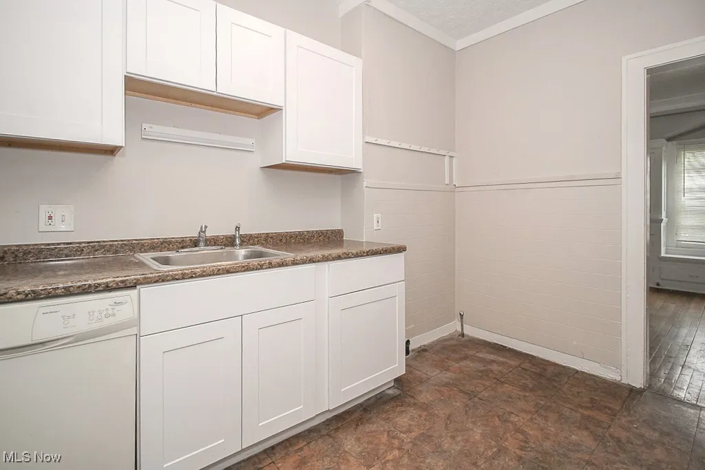 Kitchen featuring white dishwasher, dark countertops, white cabinetry, and crown molding