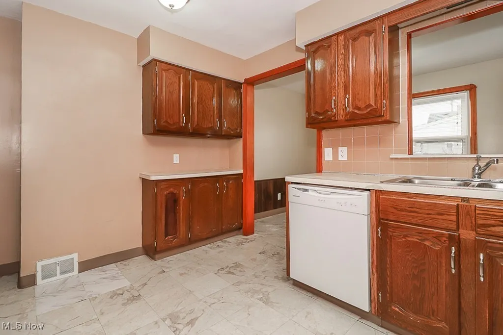 Kitchen featuring white dishwasher, light countertops, backsplash, light marble finish flooring, and brown cabinets