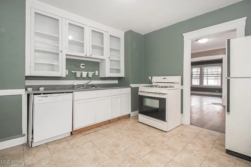 Kitchen with white appliances, glass insert cabinets, white cabinets, dark countertops, and light tile patterned floors