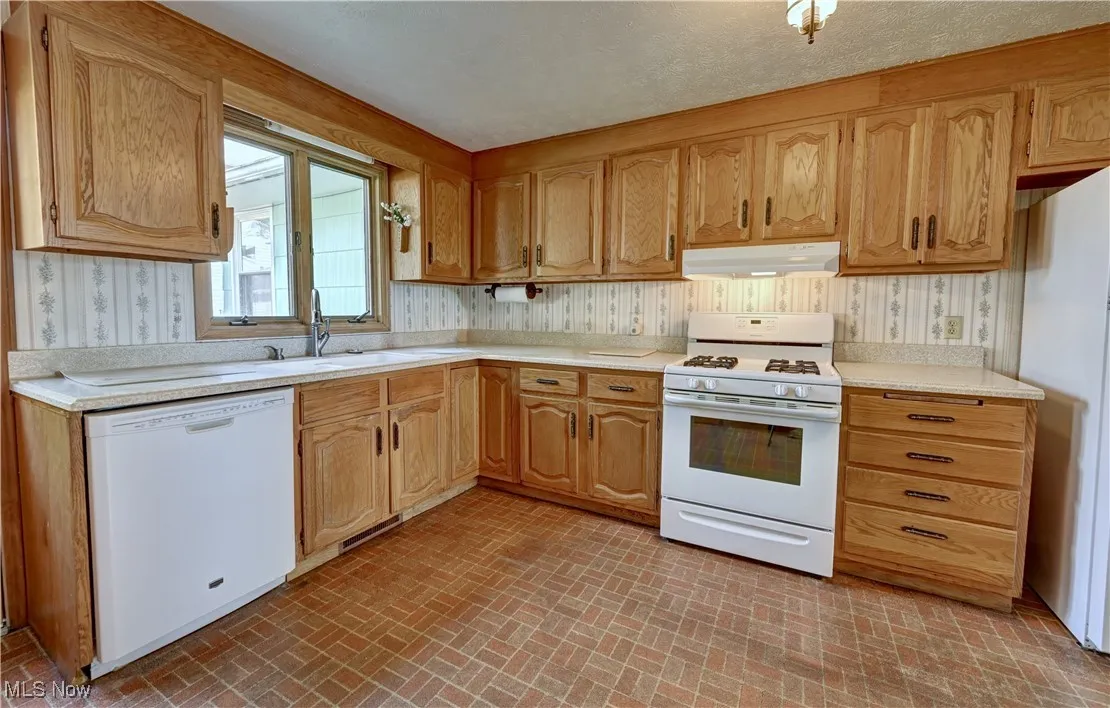 Kitchen featuring white appliances, under cabinet range hood, brick patterned flooring, light countertops, and wallpapered walls