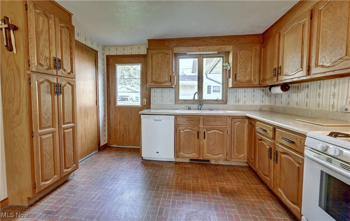 Kitchen featuring white appliances, brick patterned flooring, light countertops, wallpapered walls, and brown cabinets