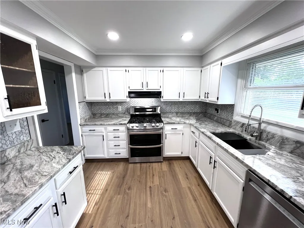 Kitchen with stainless steel appliances, under cabinet range hood, ornamental molding, white cabinetry, and light stone counters