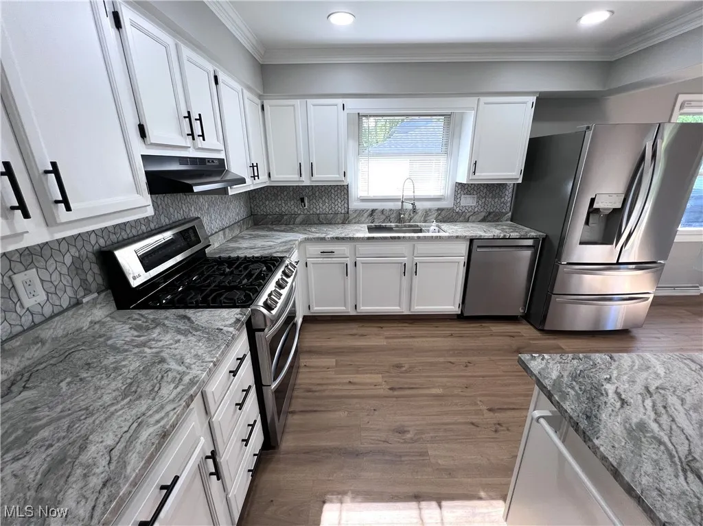Kitchen with stainless steel appliances, under cabinet range hood, crown molding, white cabinets, and light stone counters