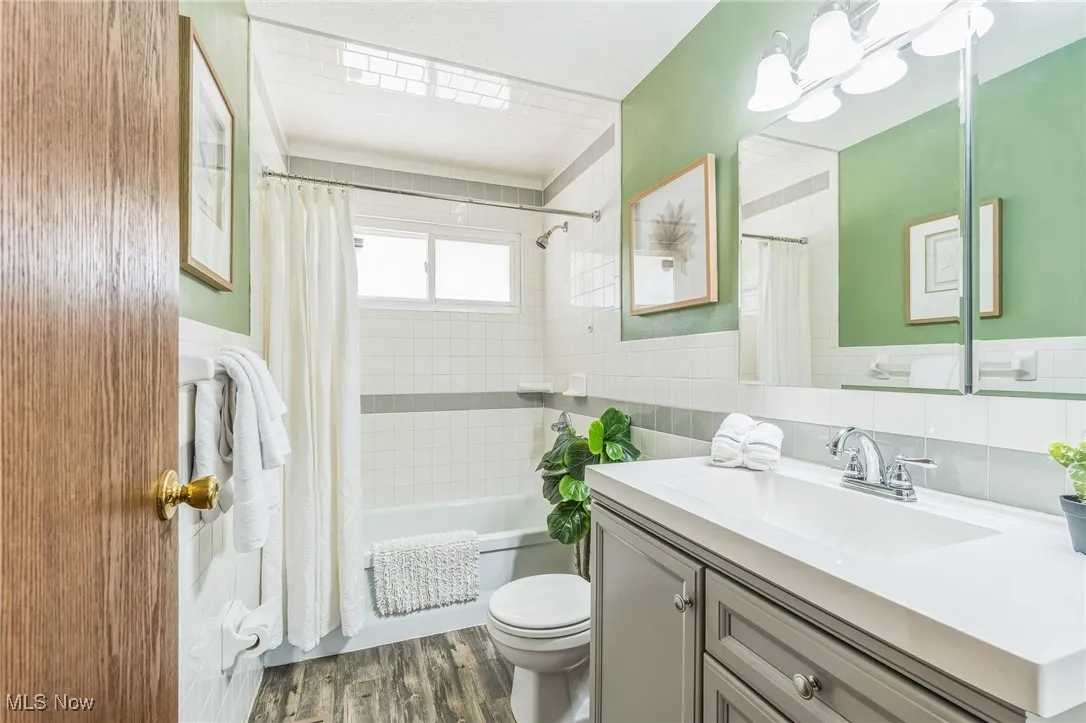 Full bathroom with tile walls, vanity, shower / bathtub combination with curtain, wood finished floors, and wainscoting