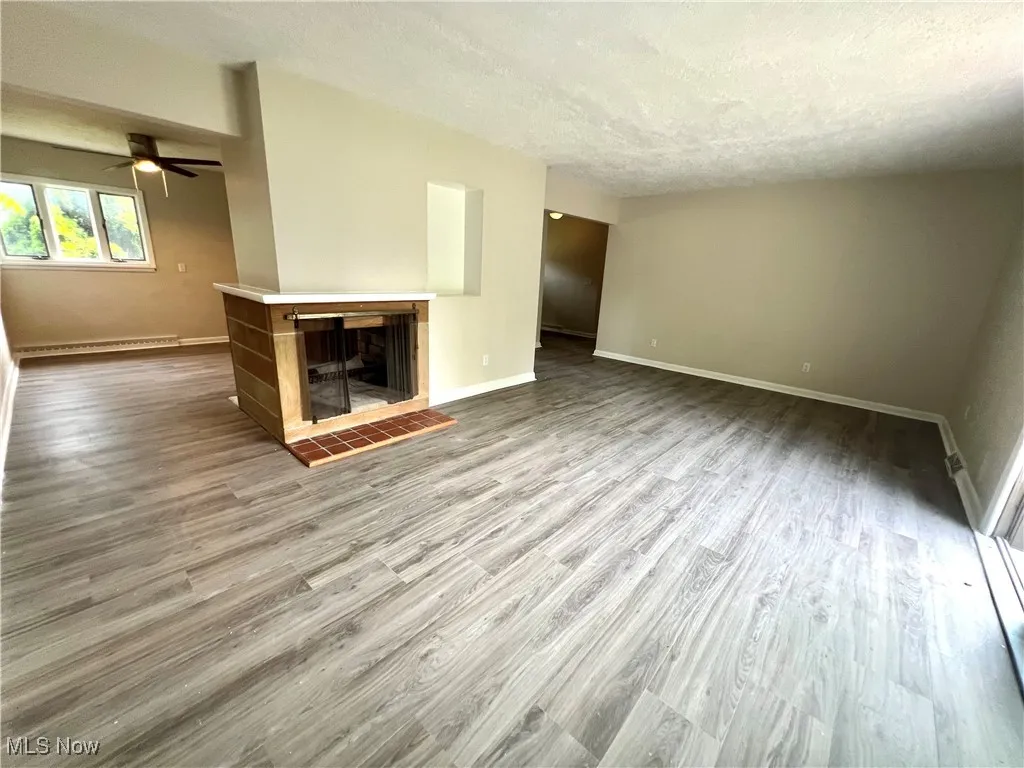 Unfurnished living room featuring wood finished floors, a textured ceiling, a tiled fireplace, a ceiling fan, and a baseboard heating unit