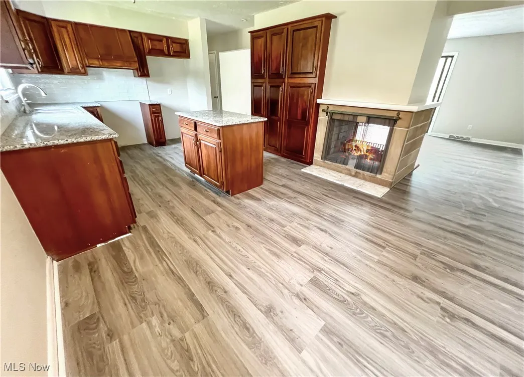 Kitchen featuring light wood finished floors, light stone countertops, a multi sided fireplace, a center island, and backsplash