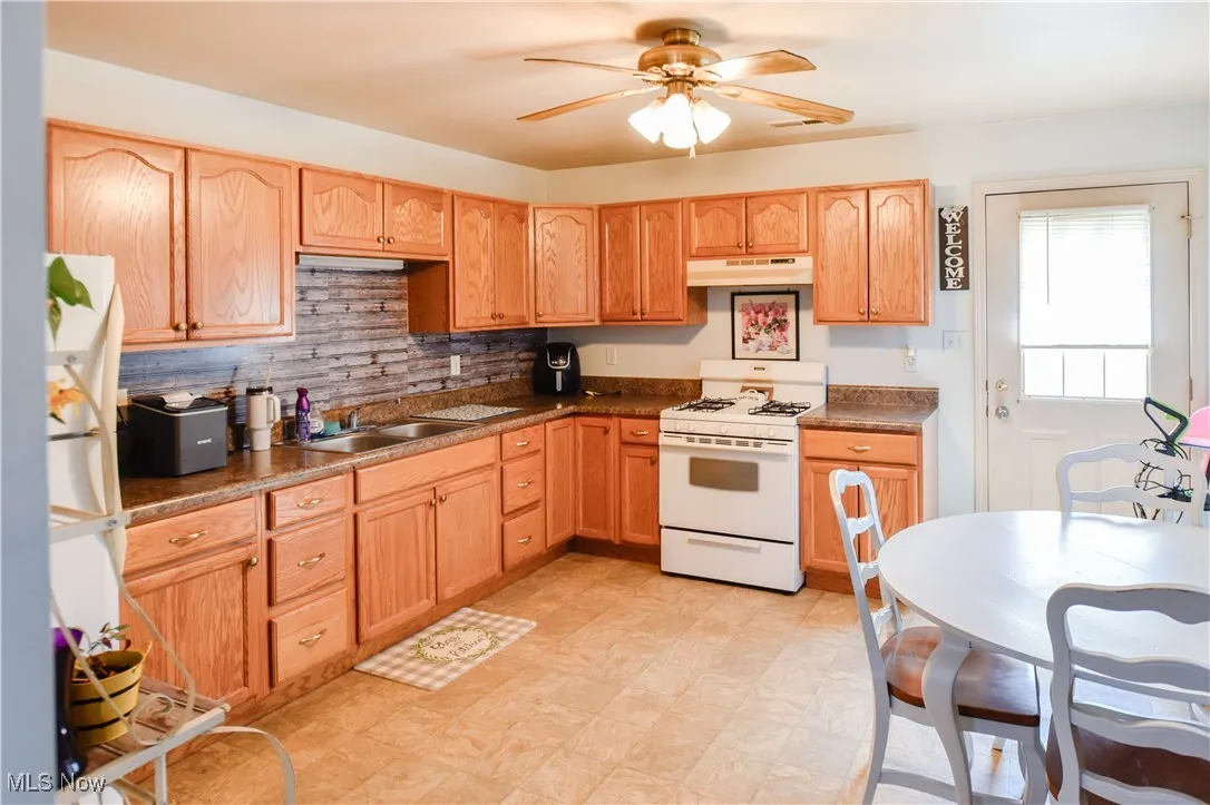 Kitchen featuring white gas stove, under cabinet range hood, ceiling fan, dark countertops, and tasteful backsplash