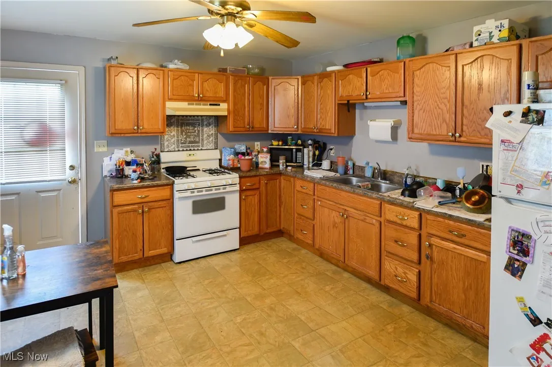 Kitchen featuring white appliances, under cabinet range hood, a ceiling fan, brown cabinets, and dark countertops