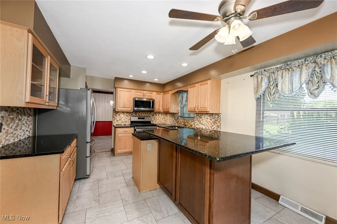 Kitchen featuring stainless steel appliances, light brown cabinetry, glass insert cabinets, ceiling fan, and backsplash