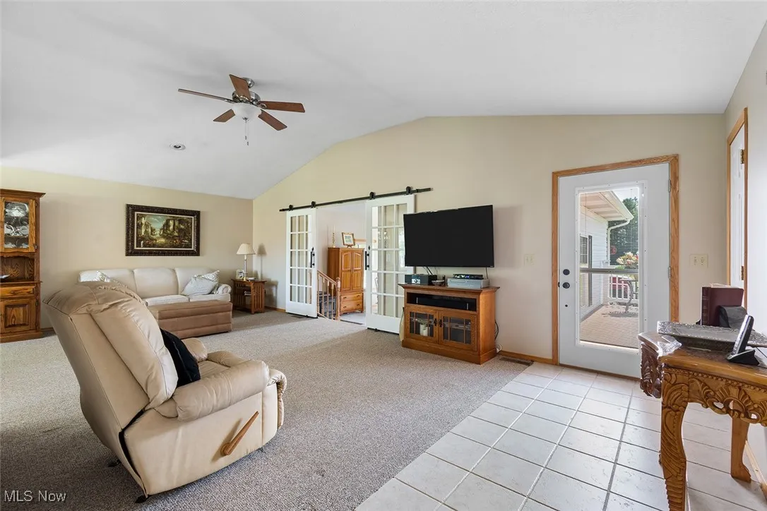 Living room with light colored carpet, vaulted ceiling, ceiling fan, light tile patterned floors, and a barn door
