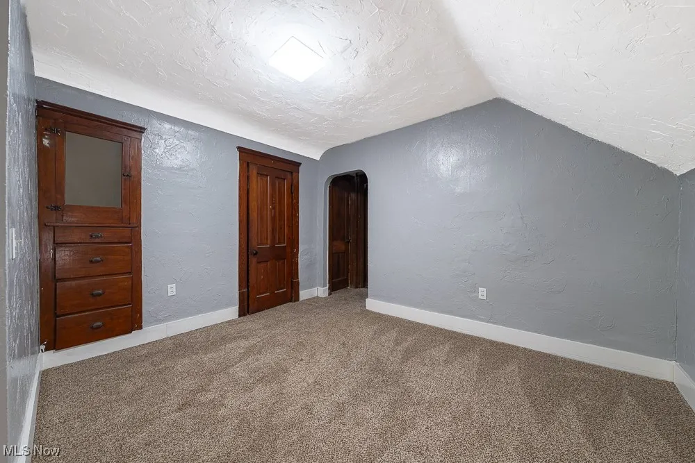 Unfurnished bedroom featuring carpet floors, vaulted ceiling, a textured ceiling, a textured wall, and arched walkways