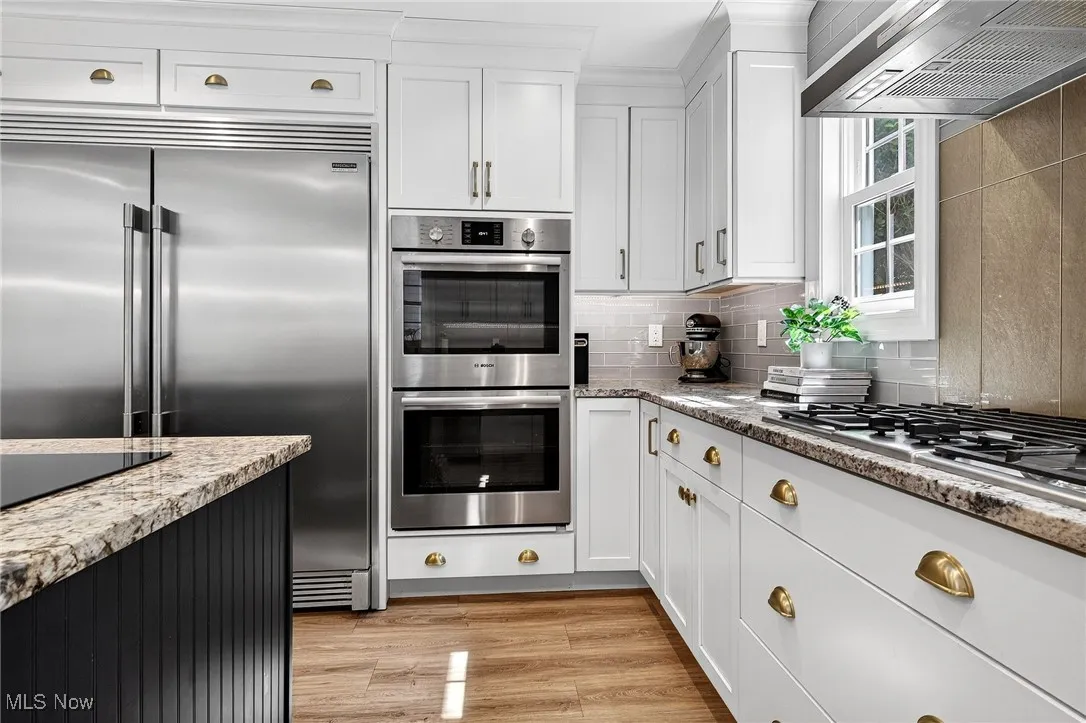Kitchen featuring stainless steel appliances, wall chimney exhaust hood, white cabinetry, backsplash, and light stone counters