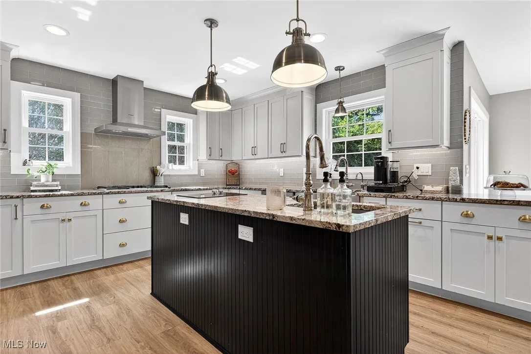 Kitchen featuring wall chimney range hood, tasteful backsplash, a kitchen island with sink, light wood-style floors, and recessed lighting