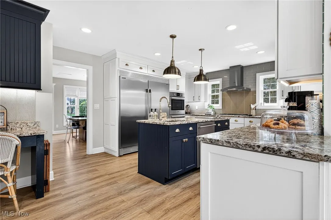 Kitchen featuring wall chimney range hood, appliances with stainless steel finishes, decorative backsplash, light wood-style flooring, and light stone counters