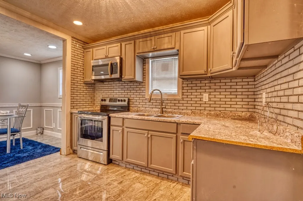 Kitchen with appliances with stainless steel finishes, a textured ceiling, crown molding, light stone countertops, and wainscoting