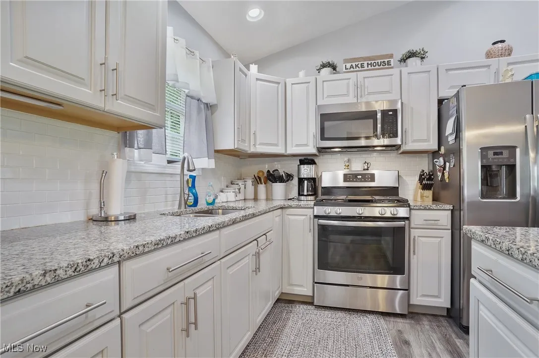 Kitchen featuring stainless steel appliances, lofted ceiling, decorative backsplash, white cabinets, and light stone counters