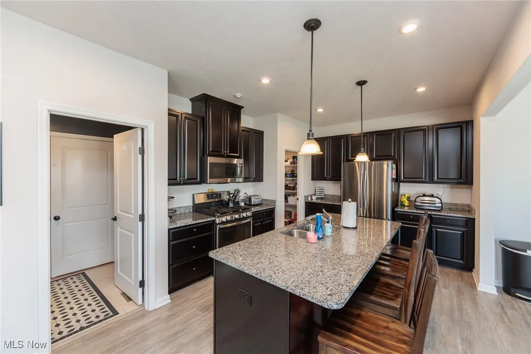 Kitchen with stainless steel appliances, an island with sink, light wood-type flooring, light stone countertops, and decorative light fixtures