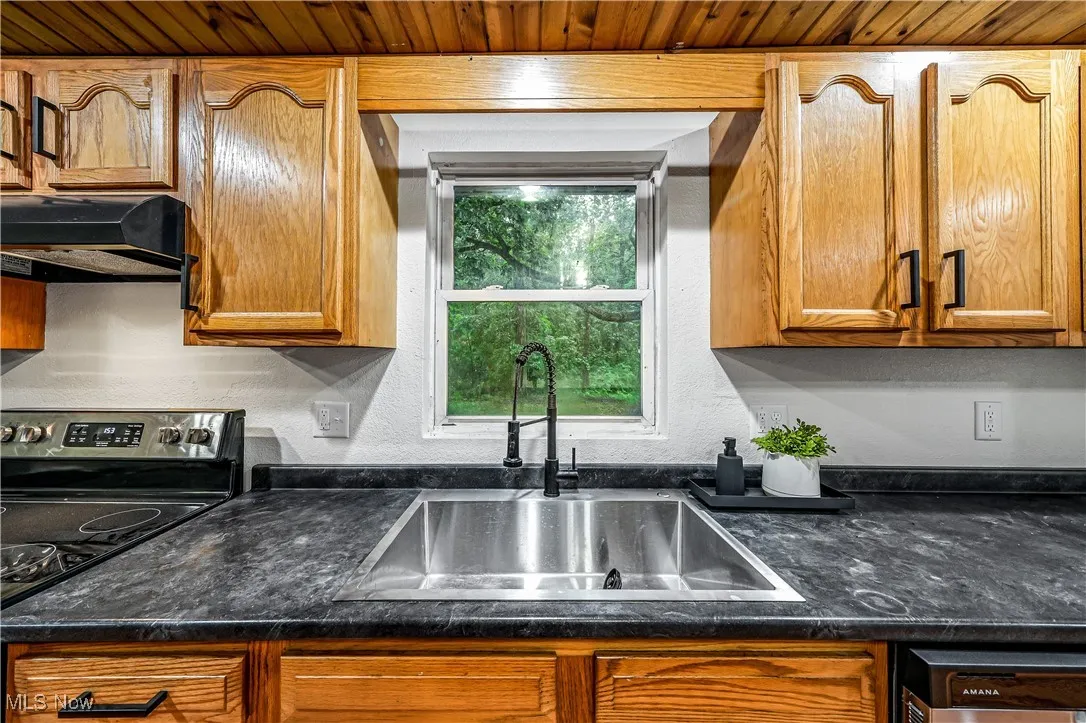 Kitchen featuring under cabinet range hood, stainless steel electric range, dark countertops, dishwasher, and brown cabinets