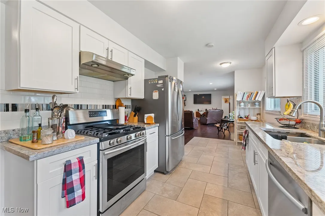 Kitchen with stainless steel appliances, under cabinet range hood, decorative backsplash, white cabinetry, and light stone countertops
