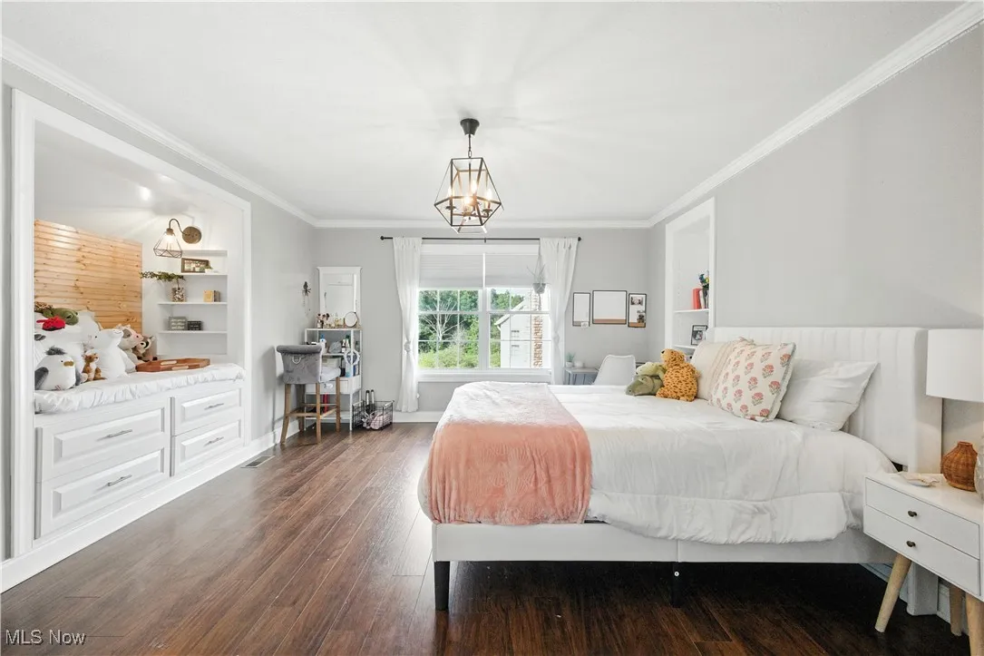 Bedroom featuring dark wood-type flooring, crown molding, and a chandelier with storage behind built-ins.