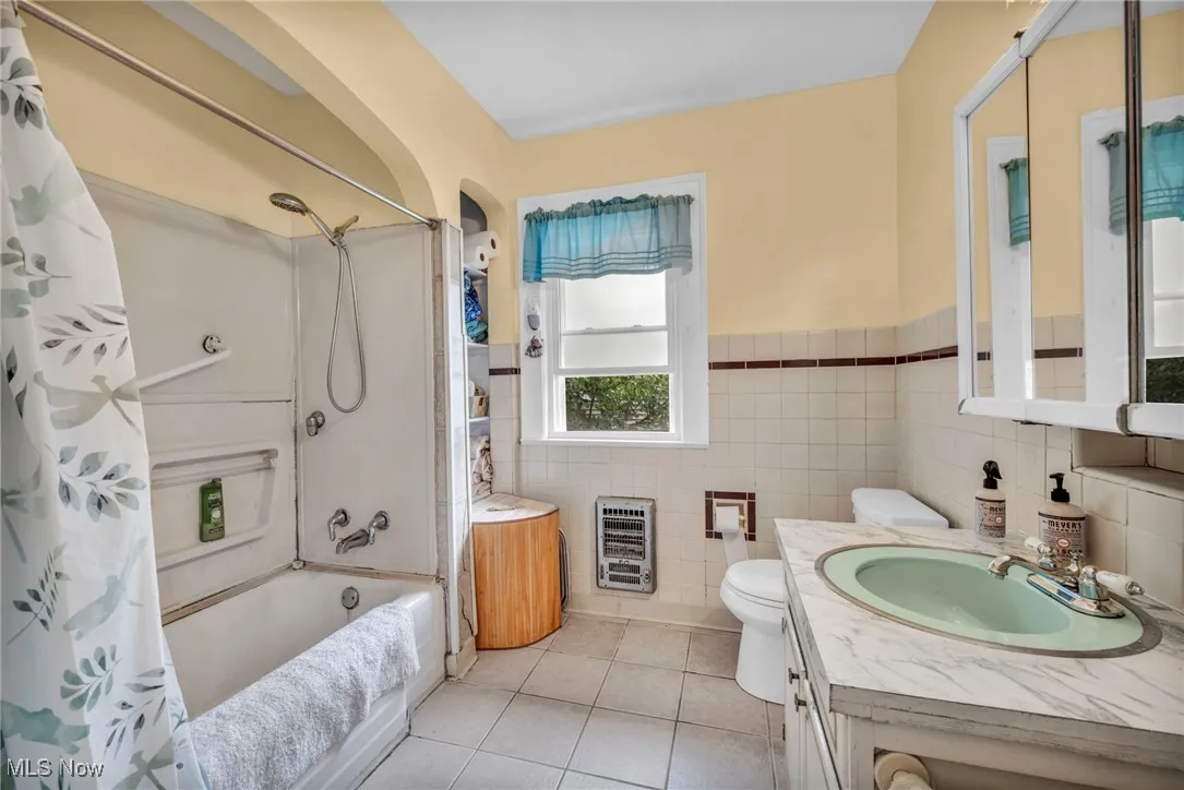 Full bathroom with heating unit, shower / bathtub combination with curtain, tile patterned floors, vanity, and tile walls