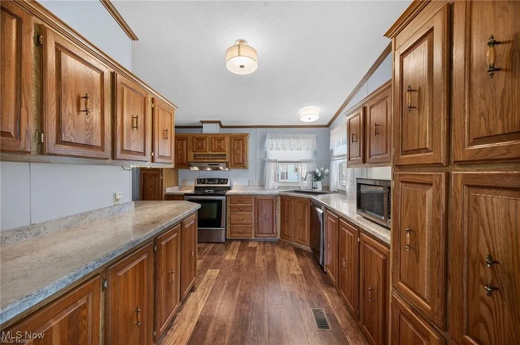 Kitchen featuring stainless steel appliances, ornamental molding, dark wood-type flooring, brown cabinetry, and light stone countertops