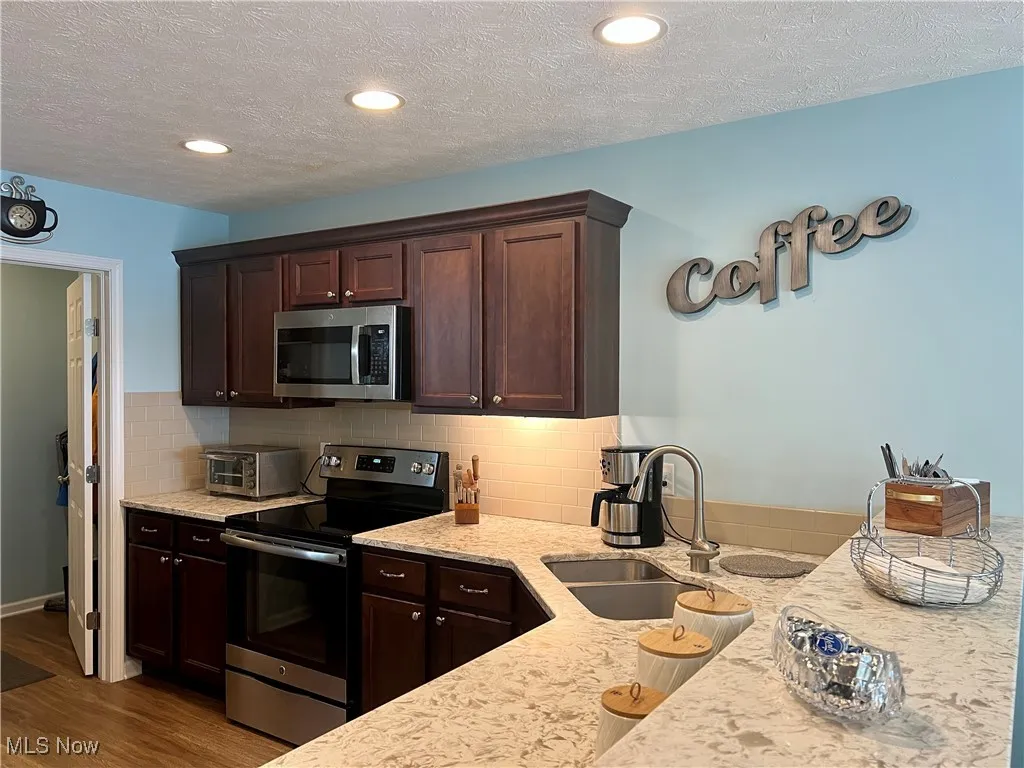 Kitchen featuring stainless steel appliances, wood finished floors, dark brown cabinets, light stone countertops, and backsplash