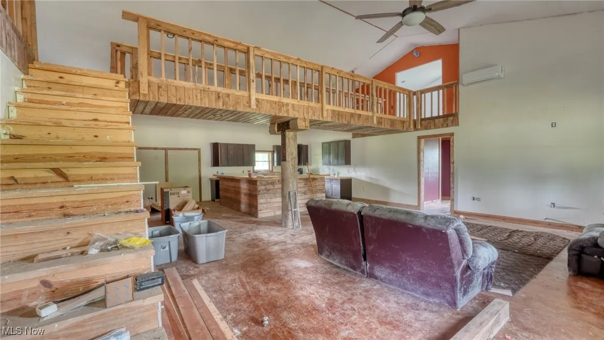 Living area with a ceiling fan, a wall unit AC, high vaulted ceiling, and stairway