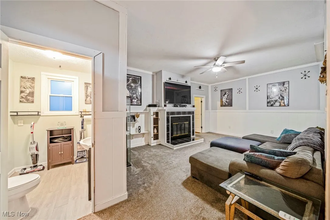 Living area featuring a ceiling fan, wainscoting, a tile fireplace, light colored carpet, and ornamental molding